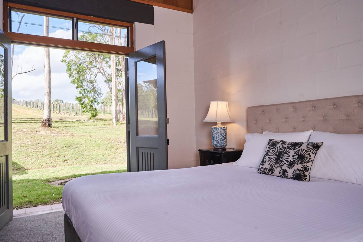 A bedroom with a large bed, a nightstand with a lamp, and a view of a grassy area through an open door.