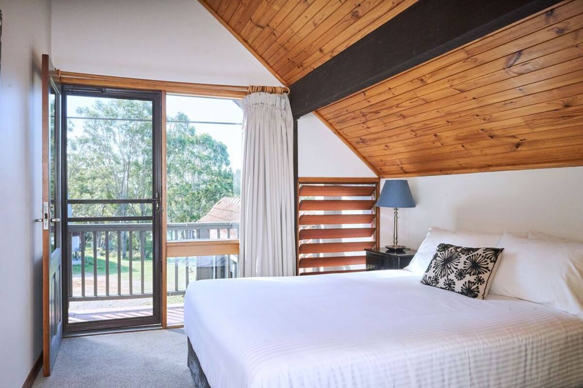 A bedroom with a large bed, a nightstand, a lamp, a window with blinds, and a view of the outdoors.