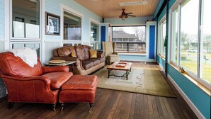 A cozy living room with a leather sofa, a coffee table, and a chair. There are ceiling fans, large windows, and a view of the outside.