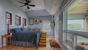 A bedroom with a bed, wooden floor, a ceiling fan, a window, and a staircase.