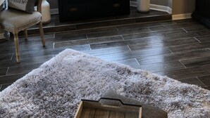 Un salon doté d’un parquet en bois foncé, d’un tapis moelleux, d’un fauteuil agrémenté d’un coussin orné d’un motif paradisiaque, d’un meuble de rangement en bois foncé et d’une lampe.