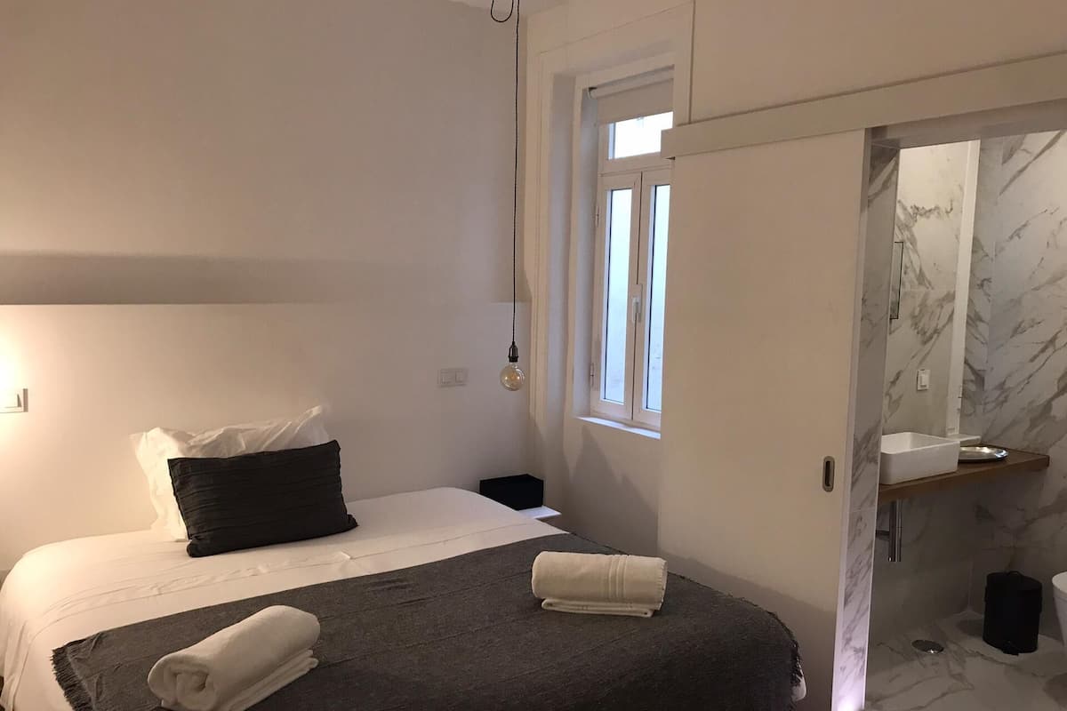 Ein Schlafzimmer mit Bett, Nachttisch, Fenster und einem Badezimmer mit Waschbecken und Spiegel.