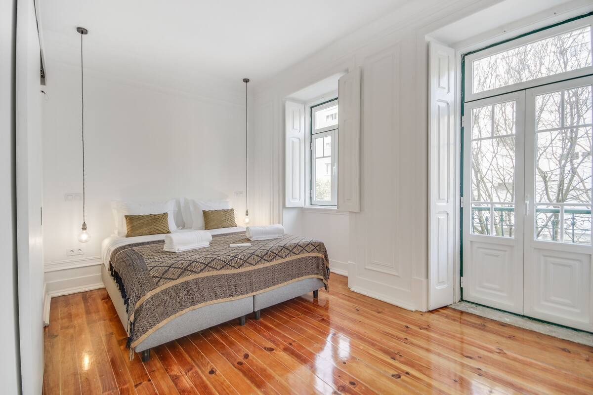 Ein Schlafzimmer mit Holzboden, einem Bett mit gemustertem Überwurf, einer Pendelleuchte und einem Fenster mit Blick auf Bäume.