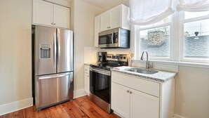 Una cocina con gabinetes blancos, electrodomésticos de acero inoxidable, encimera de granito y una ventana con cortinas blancas.