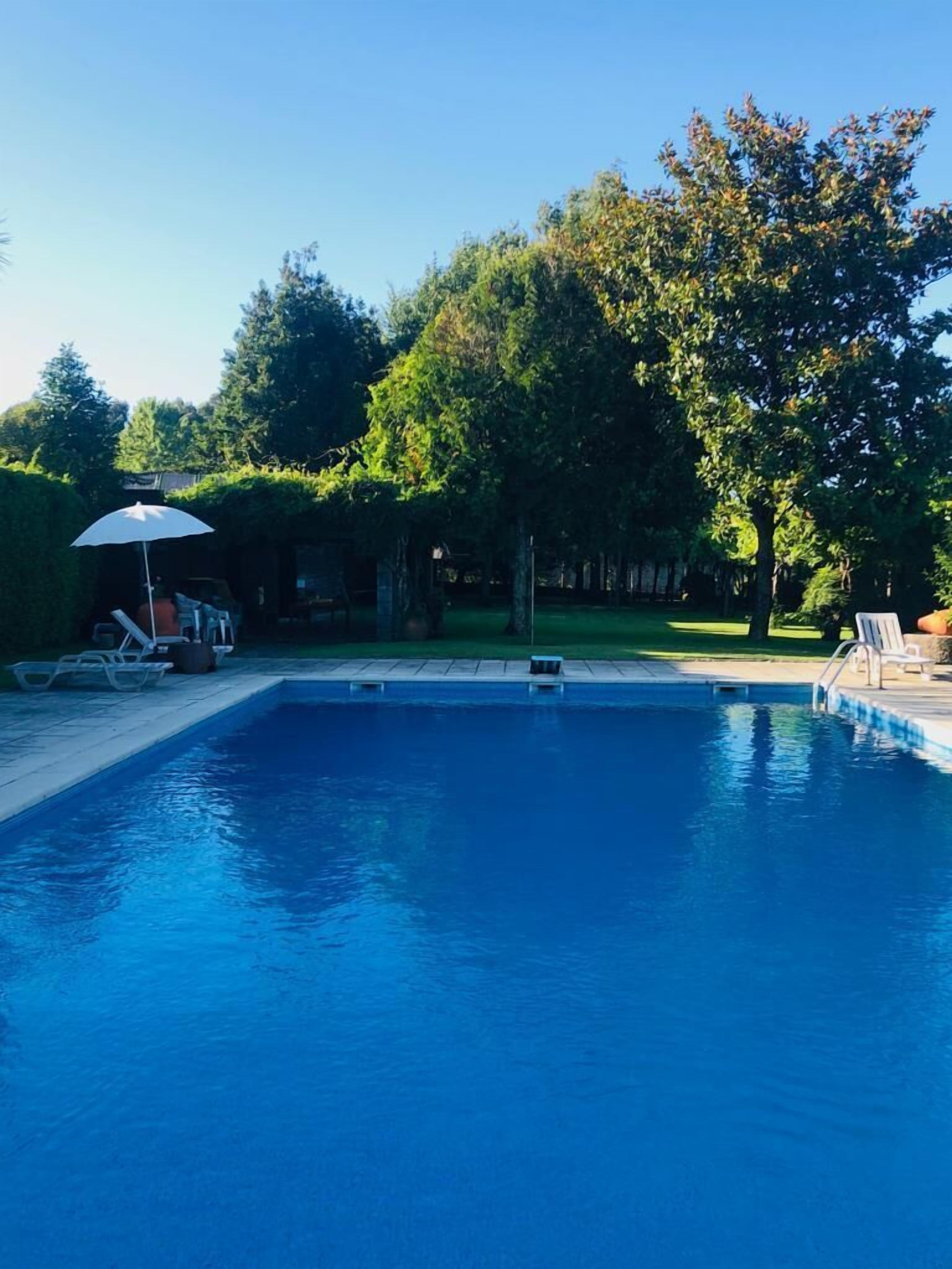 Una piscina rectangular con agua azul cristalina, rodeada por una terraza pavimentada. Hay sillones de descanso y un paraguas blanco a la izquierda.