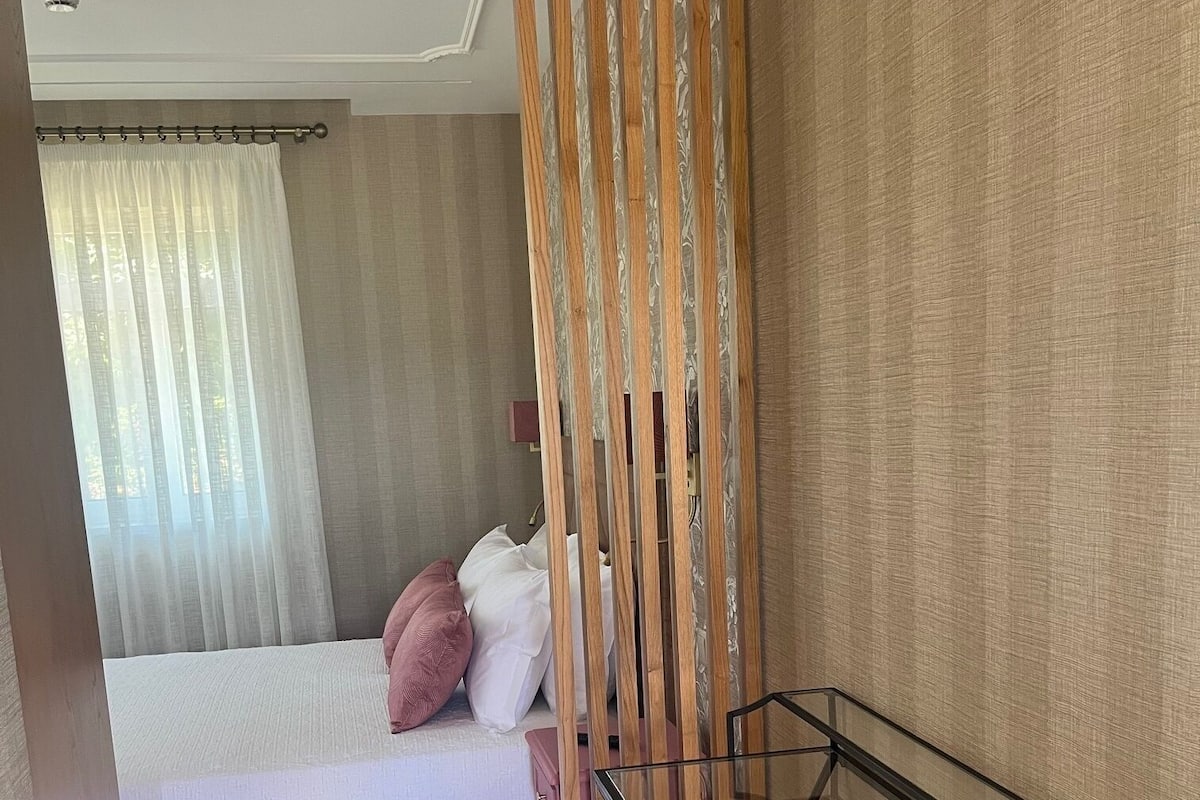 A bedroom with a bed, a wooden partition, a glass table, and a small stool.