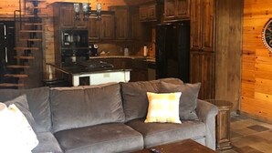 A cozy cabin interior with a sectional sofa, a wooden coffee table, and a kitchen area with wooden cabinets and a stone floor. - Relax, Fish, Visit Historic Jefferson & Caddo Lake (Jefferson)