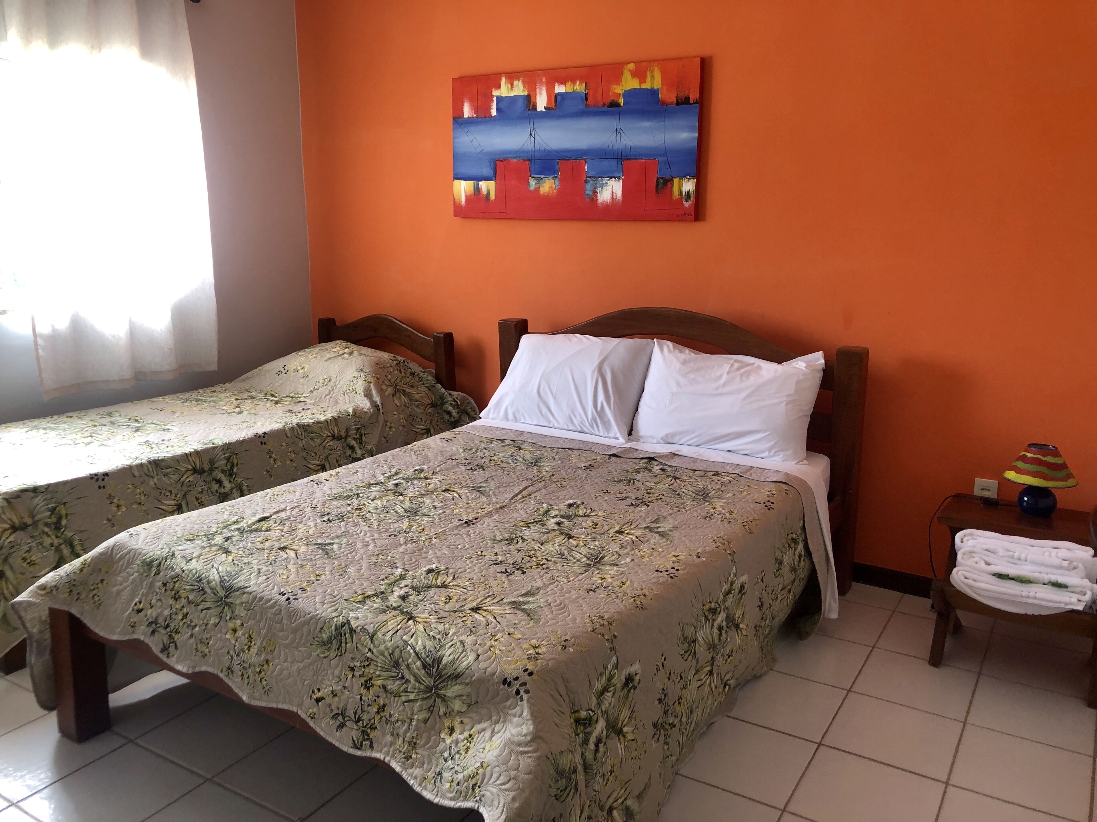 A room with two beds, each with floral bedding, a painting on the wall, and a nightstand with a lamp and towels.