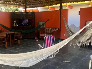 Een hangmat in een overdekte buitenruimte met een houten tafel en stoelen, een gestreepte stoel en een televisie op een plank.
