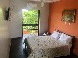 Een slaapkamer met een bed, nachtkastje, schilderij, airconditioner en raam met uitzicht op groen.
