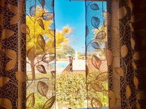 Vista a través de una ventana con cortina de estampado de hojas, una piscina y cielo despejado.