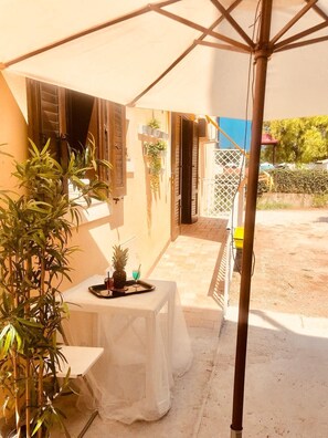 Un área de estar al aire libre techada con una mesa dispuesta para dos personas, una planta en maceta y una mesa pequeña con una bebida y una piña.