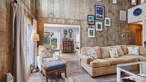 Amplia sala de estar con un sofá beige grande, un reposapiés de mimbre y una mesa de centro de vidrio. La habitación cuenta con paneles de madera en las paredes y vistas al exterior a través de un ventanal.