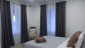 Um quarto com cama, roupa de cama cinza, duas almofadas fofas, uma cadeira e cortinas cinzas.