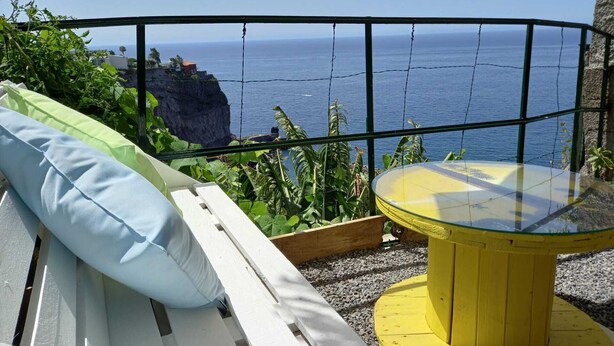 Varanda com um banco branco e mesa amarela, com vista para um penhasco e o oceano.