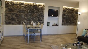 Uma área de refeições com parede de pedra ao fundo, uma TV de tela plana montada acima de uma estante, e uma mesa de centro de vidro com objetos decorativos.