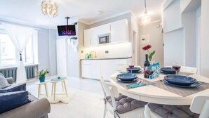 A modern dining area with a round table set for two, a flat-screen TV mounted above, and a kitchen in the background.