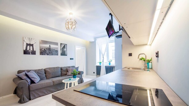 A modern living room with a grey sofa, a small white table, and a kitchen area with a black countertop and a TV mounted on the wall.
