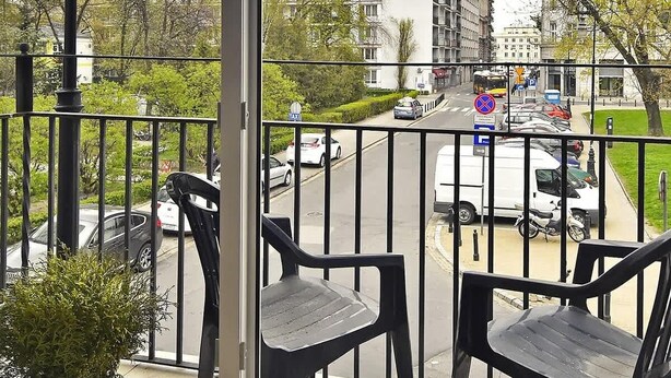 A balcony with two chairs and a potted plant, overlooking a street with buildings and parked cars.