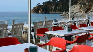 Un espacio de comedor al aire libre con mesas y sillas, con vistas a un paisaje costero con acantilados y el mar.