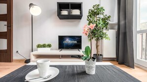 Moderni olohuone, jossa on litteänäyttöinen televisio, lattiavalaisin, ruukkukasvi sekä sohvapöytä, jonka päällä on kuppi ja aluslautanen.