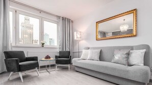 Een moderne woonkamer met een grijze bank, twee fauteuils, een kleine ronde tafel met een bloevas, een grote spiegel met gouden lijst, en een stadsgezicht door het raam.