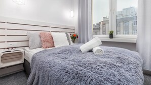 Une chambre d’hôtel moderne avec un grand lit, une table de chevet avec des verres, une petite plante et une vue sur la ville par une grande fenêtre.
