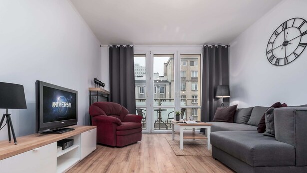 Un salon moderne avec un téléviseur à écran plat, un fauteuil rouge, un canapé gris et une grande fenêtre avec des rideaux.