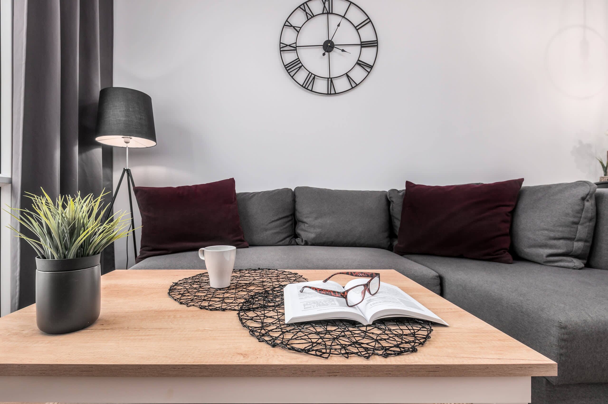 A modern living room with a grey sofa, a black clock, a floor lamp, a potted plant, a coffee cup, and an open book on a wooden table.