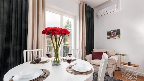A dining area with a round table set for four, a vase with red flowers, and a view of the outdoors through large windows. - Stylish Apartment | Air Conditioning | Close Center (Warszawa)