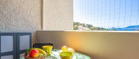 A small outdoor balcony with a tiled table, a wicker fruit basket, and a view of a coastal landscape.
