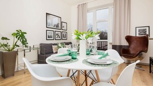 A dining area with a round table set for four, surrounded by white chairs. There are framed pictures on the wall, a potted plant, and a brown leather chair. - A Unique Apartment with a Balcony & Parking | Close to Łazienki Królewskie (Warszawa)