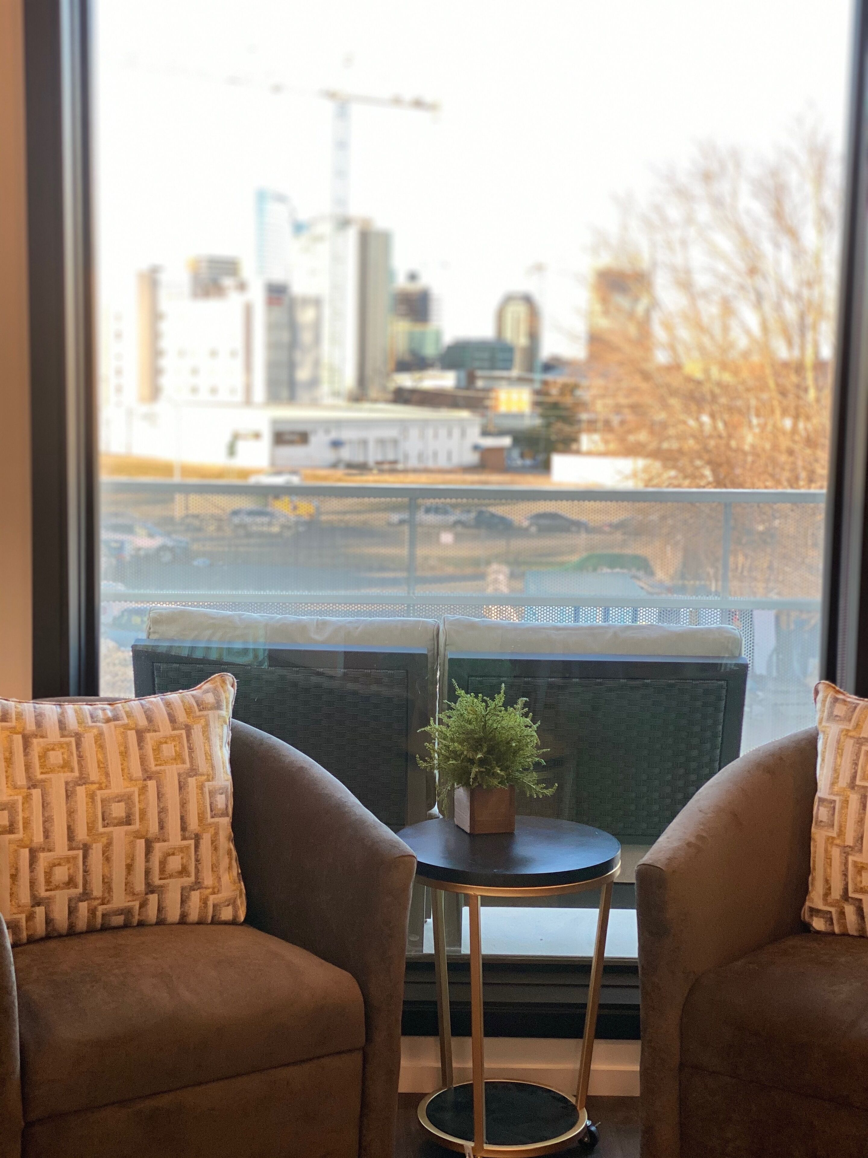 Una habitación con dos sillones marrones, una mesita redonda con una planta en maceta y vistas a un paisaje urbano a través de un ventanal grande.