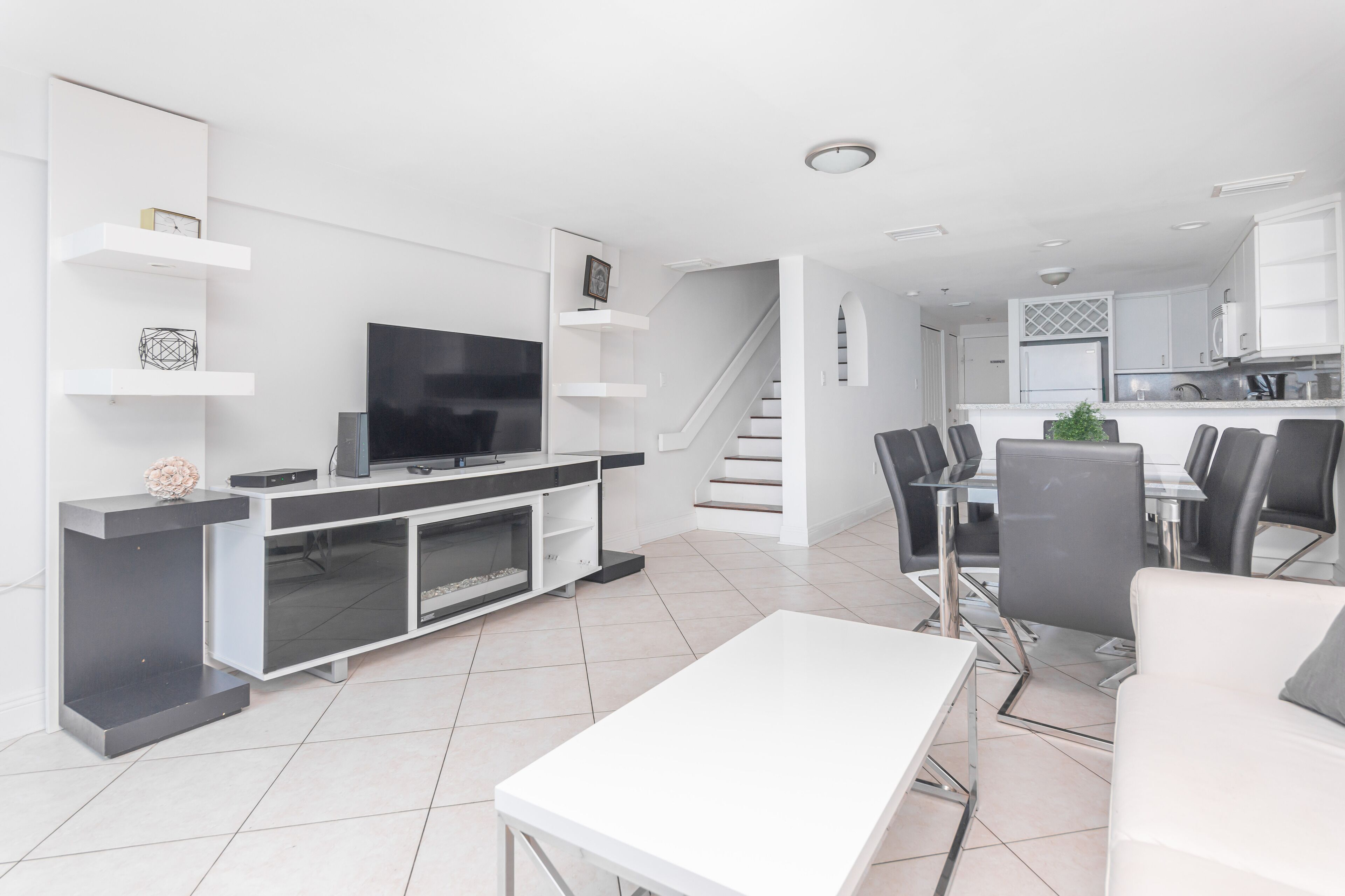 Una sala de estar moderna con una televisión de pantalla plana, una mesa de centro blanca y un comedor con sillas y una mesa.
