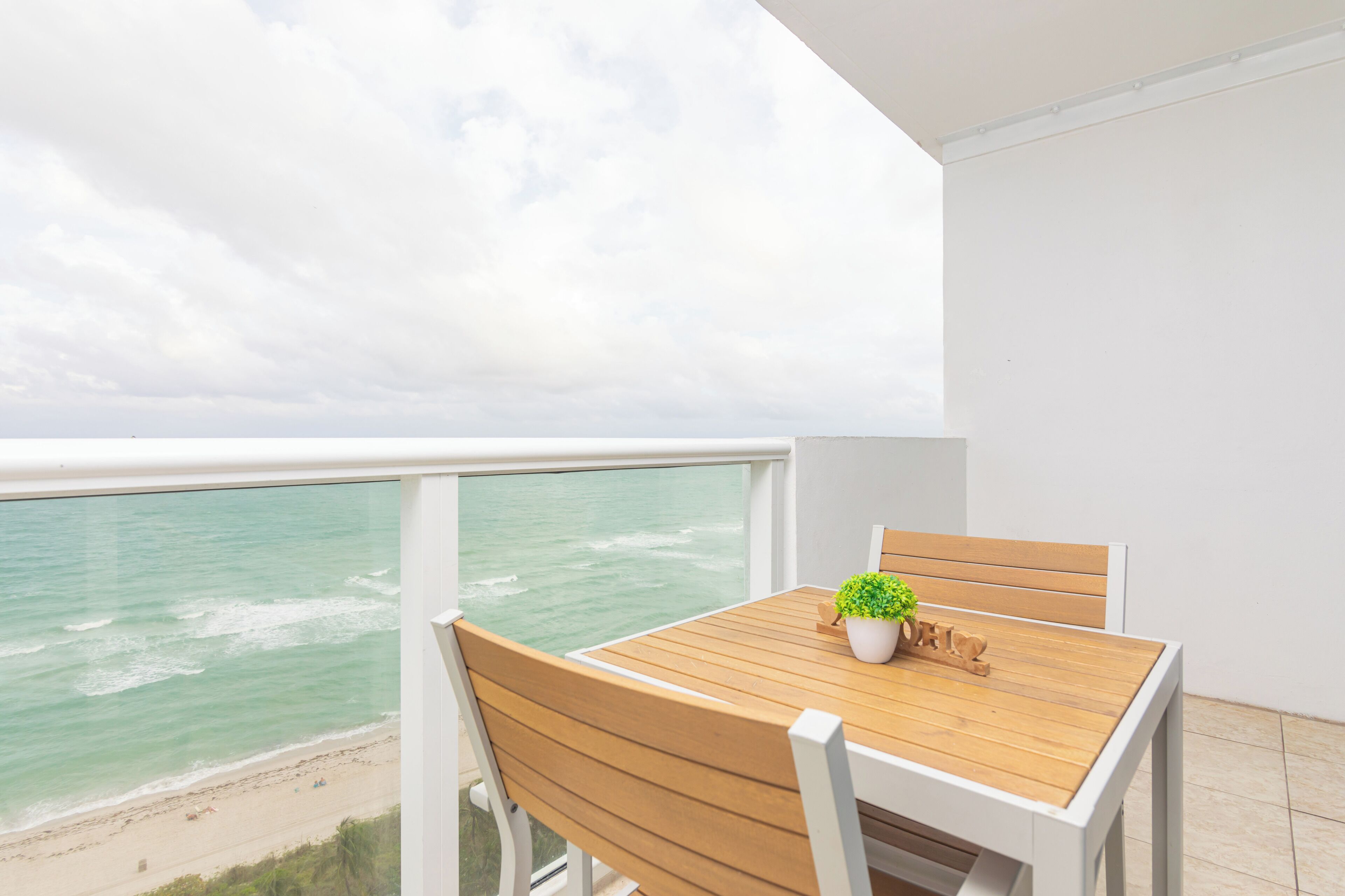 A balcony with a wooden table and chairs, a potted plant, and a view of the ocean.