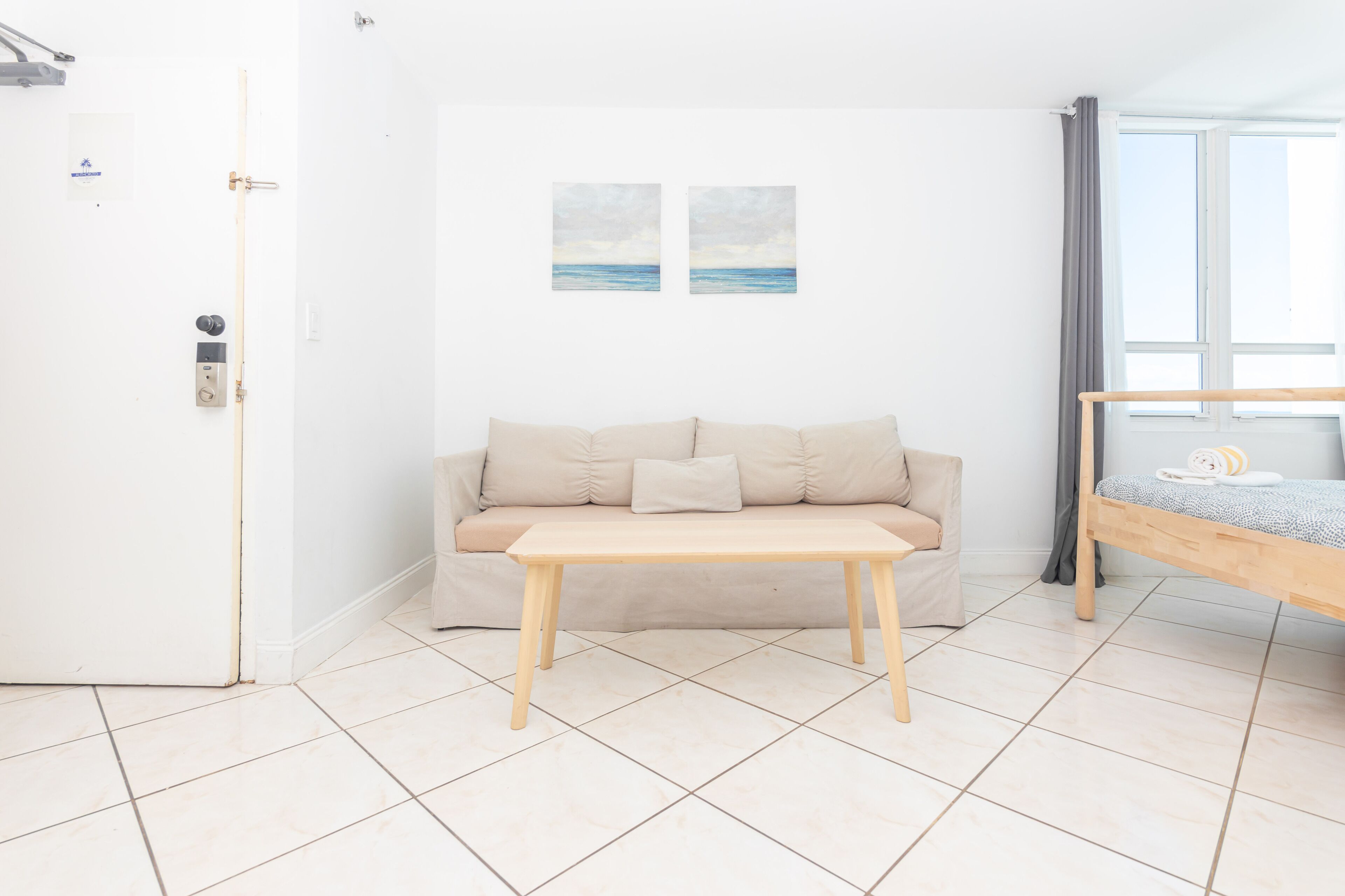 Una habitación con un sofá color beige, una mesa de madera, una cama litera con colchón azul y dos cuadros del mar en la pared.