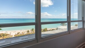 A room with a large window overlooking a beach with clear blue water and palm trees. - Girasole Apartments (Miami Beach)