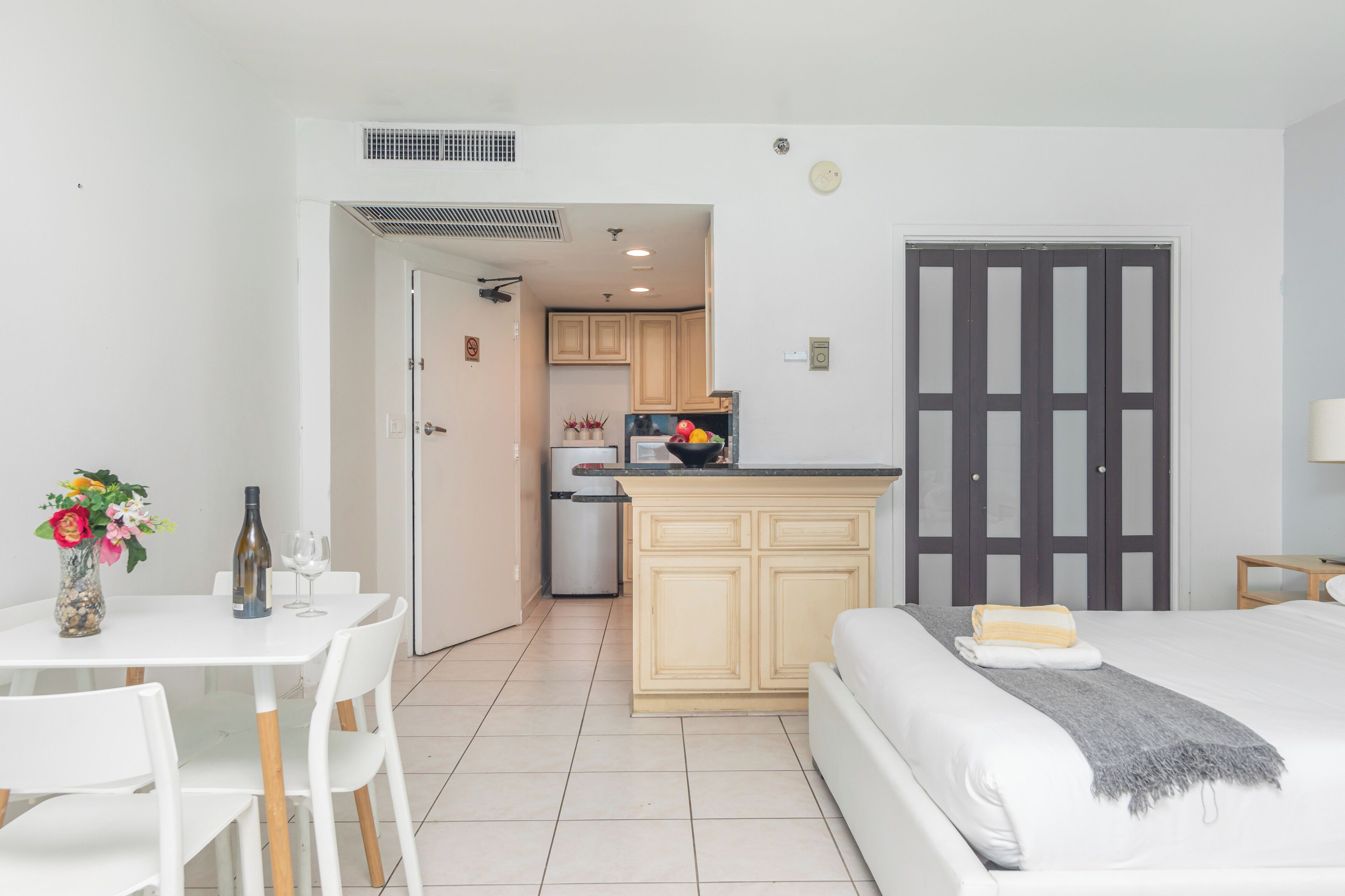 Hotelkamer met een bed, een klein tafeltje met een vaas en een fles, een kitchenette en een deur met een geëtste glazen panelen.