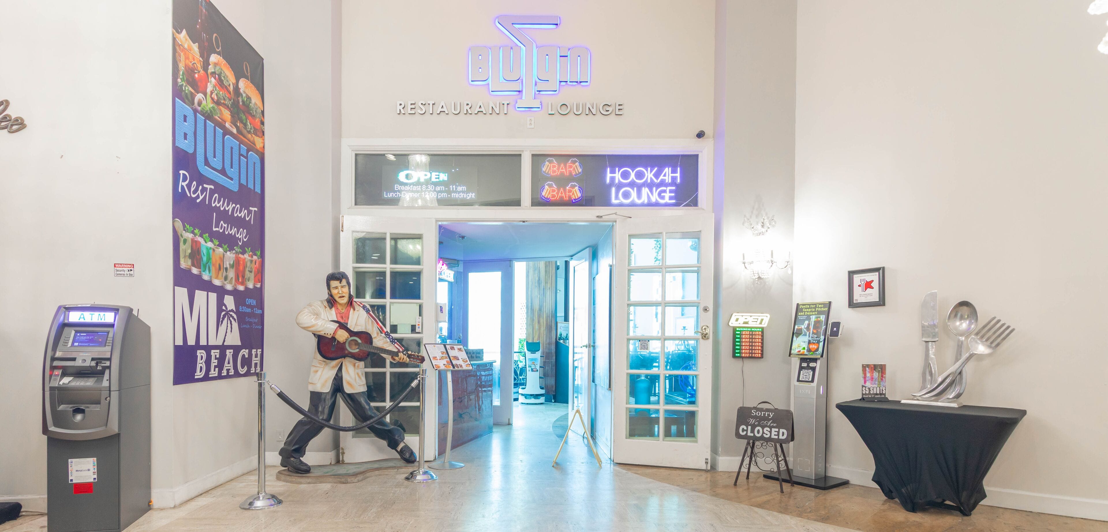 Una hall con un neon che pubblicizza un ristorante-lounge, un bancomat e una statua di un musicista.