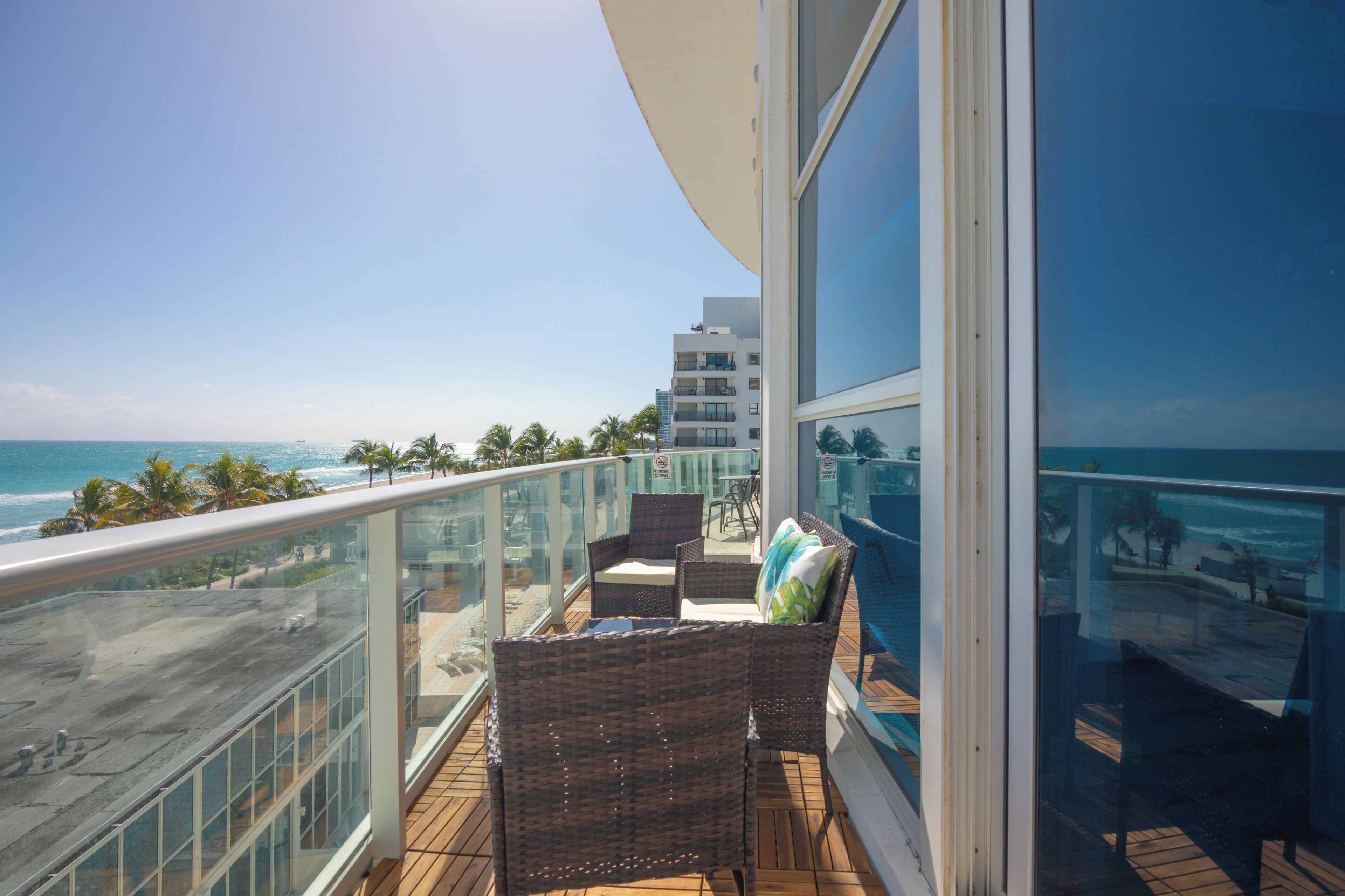 Ein Balkon mit Korbmöbeln, einer Glastrennwand und Blick auf das Meer und den Strand.