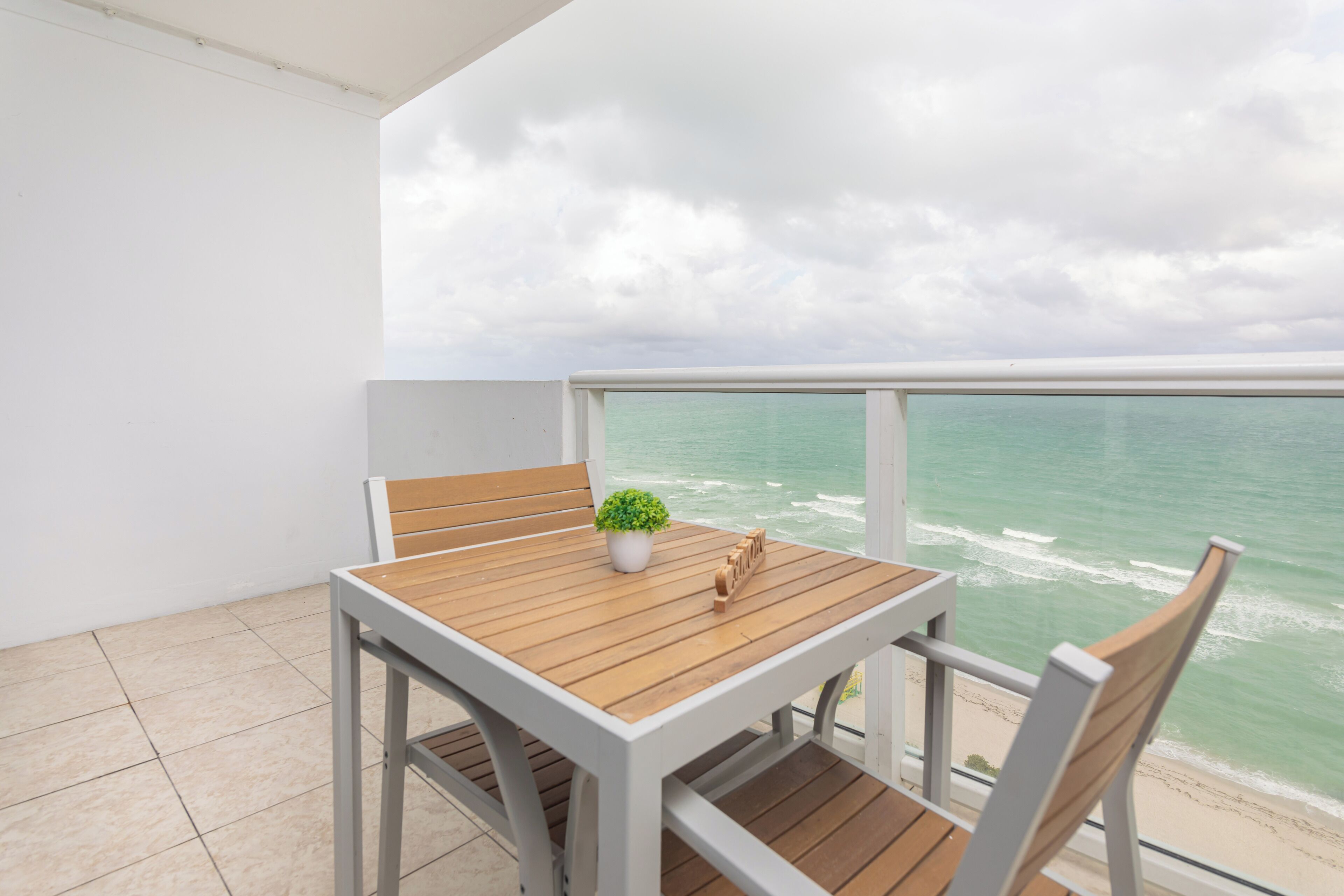 A balcony with a wooden table and chairs, a potted plant, and a view of the ocean.