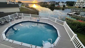 A swimming pool with a deck, surrounded by a white fence, and a sunset in the background.