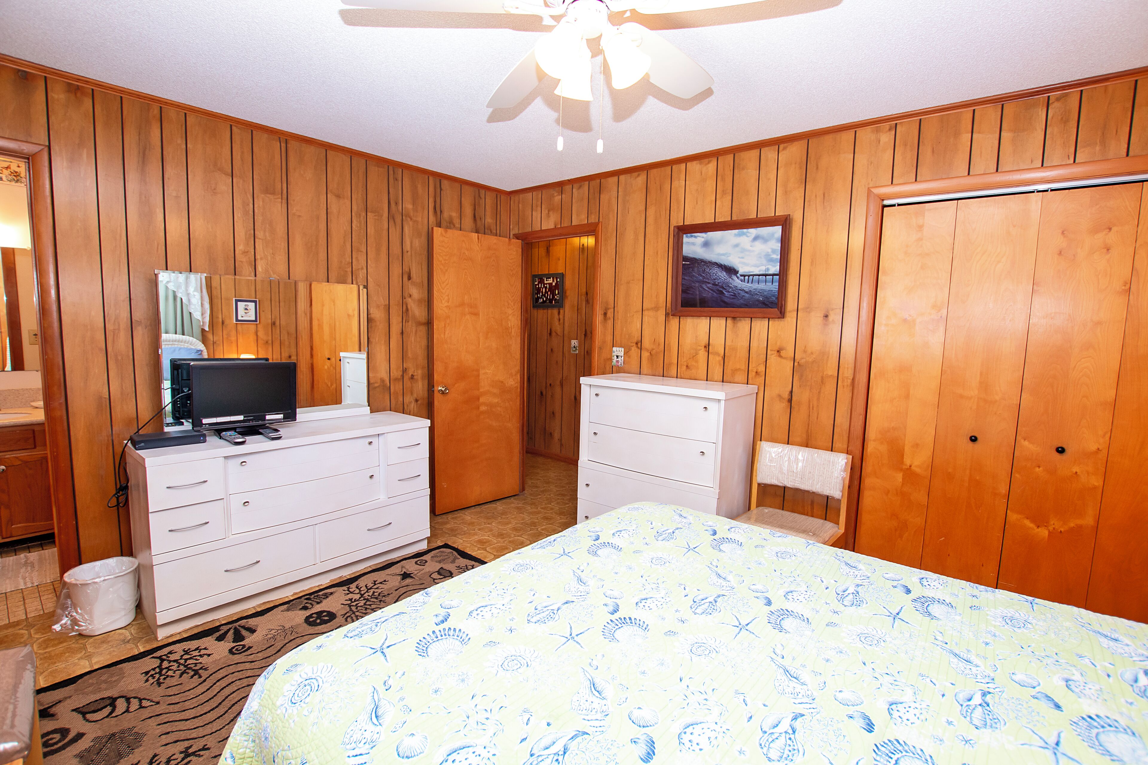 A bedroom with wooden paneling, a bed with a blue and white patterned bedspread, a white dresser, a television on the dresser, and a ceiling fan.