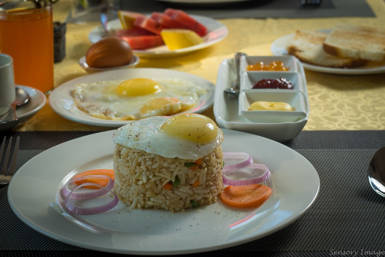 Un desayuno que incluye un plato de arroz frito coronado con un huevo frito, acompañado de cebolla y zanahoria en rodajas.