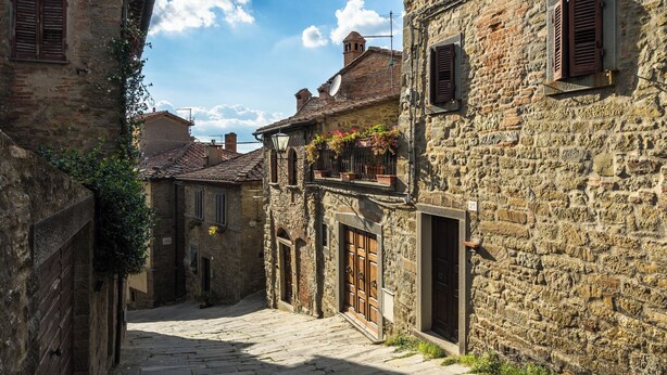 A narrow cobblestone street flanked by historic stone buildings with wooden shutters and a small balcony with hanging flowers.