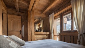 A wooden cabin bedroom with a bed, pillows, a dresser, and a window with curtains. - Cortina Lodge Stunning View R&R (Cortina d´Ampezzo)