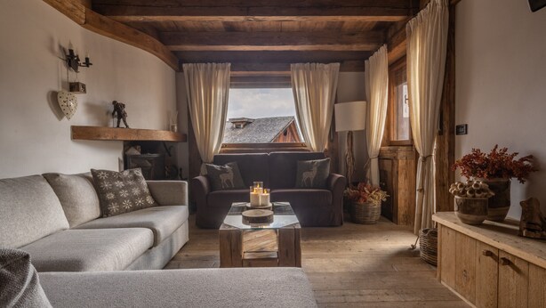 A cozy living room with a wooden ceiling, a sofa, a coffee table with candles, and a view of the outside through curtains. - Cortina Lodge Stunning View R&R (Cortina d´Ampezzo)