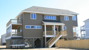 A two-story building with a balcony on the upper floor, a covered front porch, and a wooden fence in front. - Oceanside, Screened Porch Dining, Priv Pool, Yard, WW1042, Jolly Octopus (Corolla)