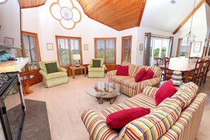 A spacious living room with a high ceiling, large windows with blinds, a variety of seating options including a striped sofa, armchairs, and a coffee table.