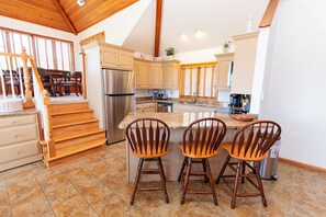 A kitchen with a central island, wooden chairs, a stainless steel refrigerator, and a staircase leading to a dining area.